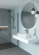 Moderne Badezimmeransicht mit einer Wanddusche, einem minimalistischen weißen Waschtisch und runden Spiegel, ergänzt durch schwarze Armaturen und LED-Beleuchtung, Ideal Standard.